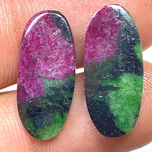 Könnte beinhalten: Zwei oval geformte Edelsteine mit polierter Oberfläche. Die Steine zeigen eine Mischung aus Farben, darunter tiefes Rosa, Grün und Schwarz. Die Edelsteine werden wahrscheinlich für Schmuck oder andere dekorative Zwecke verwendet.
