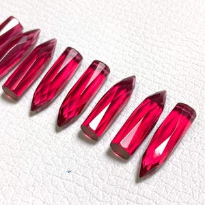 Op de afbeelding: Een verzameling gefacetteerde, langwerpige, robijnrode edelstenen. Elke edelsteen heeft een puntig uiteinde en een platte, rechthoekige basis. De edelstenen zijn diagonaal op een wit oppervlak gerangschikt, wat hun levendige kleur en ingewikkelde slijpsel benadrukt.