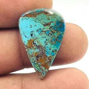 Puede incluir: Una gema pulida en forma de lágrima. La piedra presenta una mezcla de tonos turquesa, marrón y azul oscuro, con patrones y vetas detallados. La gema se sostiene entre los dedos, mostrando su tamaño y forma.
