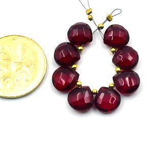 May include: A strand of faceted, deep red gemstone beads with gold-colored accents. The beads are arranged in a circular pattern. A gold coin is visible on the left side of the image, providing a size reference.