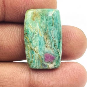 Pode incluir: Uma pedra preciosa retangular com superfície polida. A pedra apresenta uma mistura de tons verde-azulado e verde com veios castanhos e dourados. Uma pequena inclusão rosa vibrante é visível na pedra, adicionando um toque de cor.