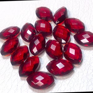 May include: A collection of faceted, oval-shaped, deep red gemstones. The gems are arranged on a white surface, each displaying multiple reflective facets. The color is a rich, saturated red, suggesting a high-quality material. These could be used for jewelry making.