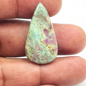 Pode incluir: Uma pedra preciosa em forma de gota. A pedra apresenta uma base verde clara com manchas rosa, roxas e amarelas. A superfície polida é lisa e reflete a luz. Ideal para joalharia.