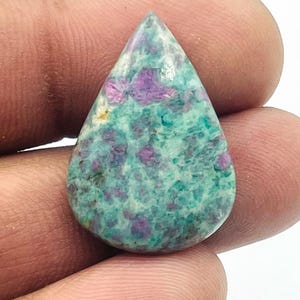 Pode incluir: Uma pedra preciosa polida em forma de gota. A pedra apresenta um padrão salpicado de tons turquesa e roxo. A pedra preciosa é segurada em uma mão, destacando sua forma. A pedra provavelmente se destina a joias ou fins decorativos.