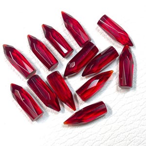 May include: A collection of faceted, deep red gemstones. Each gem is elongated with a pointed tip and a geometric cut, showcasing internal light refraction. The gems are scattered on a white surface, highlighting their rich color and clarity.