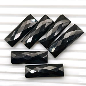 Könnte beinhalten: Sechs facettierte, rechteckige schwarze Edelsteine, angeordnet auf einer weißen Oberfläche. Die Edelsteine haben ein geometrisches Muster aus abgewinkelten Schnitten, wodurch eine reflektierende Oberfläche entsteht. Die Edelsteine sind etwa 2,5 cm lang.