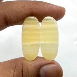 May include: Two oval-shaped, polished gemstones with a translucent, banded appearance. The stones have a pale yellow colour with lighter, creamy-white bands running horizontally. The gemstones are held between fingers against a white background.