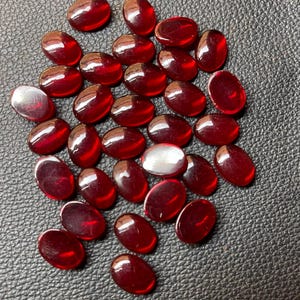 May include: A collection of oval-shaped, polished red glass beads. Some beads have a reflective, silver-like appearance. The beads are scattered on a textured, dark gray surface, creating a visually appealing arrangement.