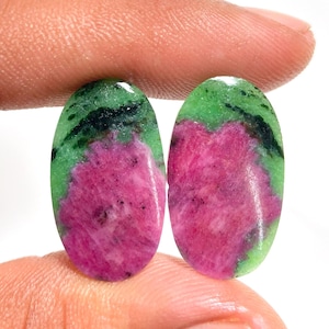 May include: Two oval-shaped gemstones. Each stone features a vibrant pink center with green and black accents. The stones are held between fingers, showcasing their polished surfaces and natural color variations. These gemstones are ideal for jewelry making.