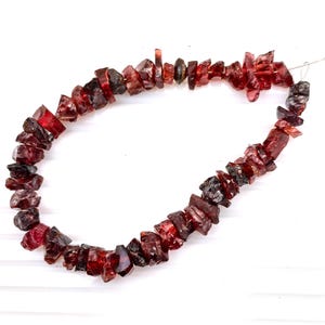 Könnte beinhalten: Eine Halskette aus grob geschnittenen, dunkelroten Granatsteinen. Die Steine variieren in Größe und Form und sind auf einem dünnen, silberfarbenen Draht aufgereiht. Die Halskette wird vor einem weißen Hintergrund gezeigt.