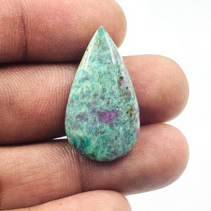 Pode incluir: Uma pedra preciosa em forma de gota com um padrão manchado de tons verde-azulado, verdes e magenta. A pedra polida é segurada entre os dedos, mostrando suas cores e texturas naturais. A gema é provavelmente usada para joias ou fins decorativos.
