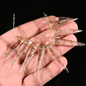 May include: A strand of seven clear, triangular, spike-shaped beads featuring internal copper-colored glitter inclusions. Each bead is separated by a small, round, metallic gold-toned spacer bead on a thin, flexible, silver-colored wire string.