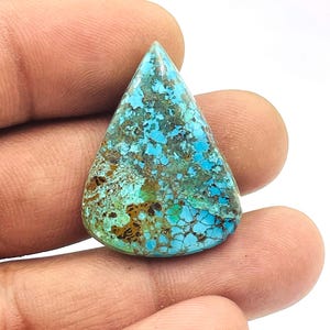Op de afbeelding: Een traanvormige turkoois edelsteen met een gevlekt patroon van blauwe, bruine en groene tinten. De gepolijste steen wordt in een hand gehouden en toont zijn natuurlijke kleurvariaties en unieke textuur. De edelsteen is een levendig voorbeeld van natuursteen.