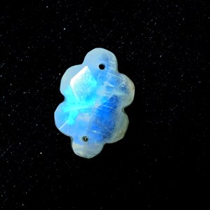 May include: A flower-shaped gemstone with a blue and white colour gradient. The stone has a faceted surface and two small holes. The gemstone is set against a black background.