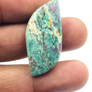 Pode incluir: Uma pedra preciosa polida com uma forma curva e irregular. A pedra apresenta uma mistura de cores, incluindo verde-azulado, branco e manchas de roxo e dourado. A pedra preciosa é segurada entre dois dedos.