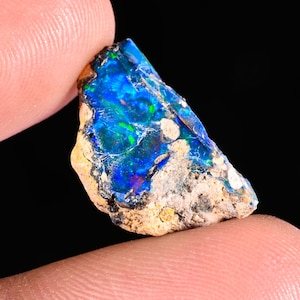 Puede incluir: Una vibrante gema de ópalo en bruto con una llamativa exhibición de iridiscencia azul, verde y roja. La piedra en bruto tiene una matriz beige texturizada y se sostiene entre los dedos sobre un fondo negro. Esta gema natural es ideal para la fabricación de joyas.