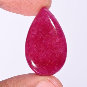 May include: A teardrop-shaped, polished gemstone in a vibrant magenta hue. The stone has a smooth, reflective surface with subtle internal textures. The gem is held between two fingers, showcasing its size and shape. This gemstone is suitable for jewelry making.