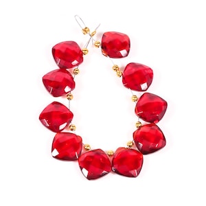 May include: Strand of faceted, square-shaped, deep red beads. The beads are strung on a thin, silver-colored wire with small gold-colored beads between each red bead. The beads are arranged in a curved line against a white background.
