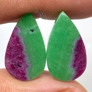 Könnte beinhalten: Zwei tropfenförmige Edelsteine. Jeder Edelstein hat eine leuchtend grüne obere Hälfte und eine tiefmagentafarbene untere Hälfte. Die Edelsteine werden zwischen den Fingern gehalten, wodurch ihre polierten Oberflächen und natürlichen Farbvariationen zur Geltung kommen.
