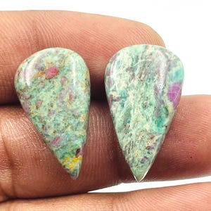Pode incluir: Duas pedras preciosas em forma de gota, com uma mistura de tons verdes, rosa e vermelhos. As pedras têm uma superfície polida e lisa e são seguradas em uma mão. As pedras preciosas são provavelmente para fazer joias ou colecionar.