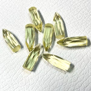 May include: A collection of faceted, light yellow gemstones, each cut into a bullet shape. The gems are arranged on a white surface, showcasing their clarity and the play of light across their many facets.