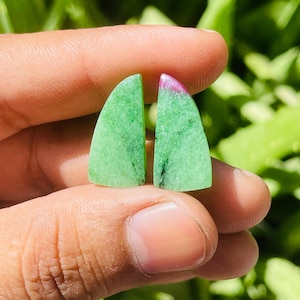 May include: Two polished ruby zoisite gemstones. Each stone is a vibrant green with black inclusions and a pinkish-red top. The gemstones are held between fingers against a blurred green background.