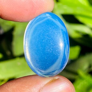 Peut inclure: Une pierre précieuse ovale polie, de couleur bleue, avec des veines blanches. La pierre est tenue entre deux doigts, mettant en valeur sa surface lisse et réfléchissante. Le fond est un feuillage vert flou, accentuant la couleur et la forme de la pierre.
