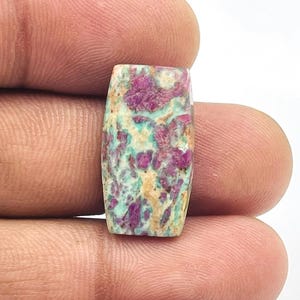 Pode incluir: Uma pedra preciosa retangular com uma superfície polida. A pedra apresenta uma mistura de cores, incluindo tons de roxo, verde-azulado e bege. O padrão é uma mistura de redemoinhos e manchas. A pedra preciosa é segurada entre os dedos.