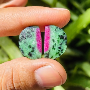 May include: Two polished zoisite gemstones, each with a half-moon shape. The stones feature a vibrant pink stripe through the center, surrounded by a speckled green and black pattern. The gemstones are held between fingers.