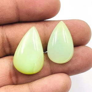 May include: Two pear-shaped, pale green gemstones. The stones have a smooth, polished surface and a slight translucence. The gemstones are displayed against a neutral background.