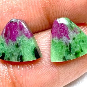 Puede incluir: Dos piedras preciosas triangulares con una vibrante combinación de colores. La parte superior de cada piedra presenta un tono magenta intenso, que se transforma en una base verde con motas negras. Las piedras están pulidas y lisas.