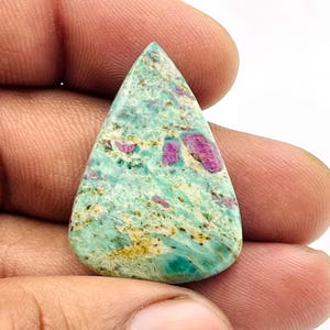 Pode incluir: Uma pedra preciosa em forma de gota com uma mistura de cores. A pedra apresenta uma base verde clara com manchas amarelas e pontos escuros. Inclusões rosa brilhante e magenta estão espalhadas por toda a pedra.