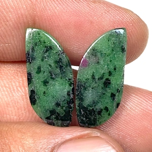 May include: Two polished, teardrop-shaped gemstones. The stones are a deep green with black inclusions and a few small red spots. The gems are held in a person's hand, showcasing their natural beauty and unique patterns. These stones are ideal for jewelry making.