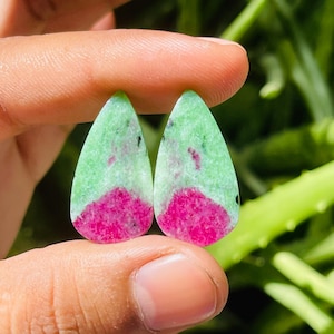 May include: Two teardrop-shaped gemstones with a vibrant color scheme. The top portion is a light green, transitioning to a bright pink at the bottom. Small black specks are scattered throughout. The gemstones are held between fingers.