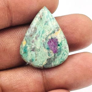 Pode incluir: Uma pedra preciosa em forma de gota com uma mistura de tons verdes, roxos e amarelos. A pedra tem uma aparência manchada, com as cores se misturando em um padrão natural. A pedra preciosa é segurada em uma mão.