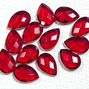 May include: A collection of faceted, teardrop-shaped red gemstones. The gems are arranged on a white surface, each displaying a brilliant, reflective quality. The cut of the stones creates multiple facets, enhancing their sparkle and depth. These gems are suitable for jewelry making.