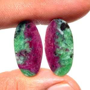 May include: Two oval-shaped ruby zoisite gemstones. The gemstones display a vibrant mix of colours, with a prominent magenta hue complemented by patches of green and black speckles. The stones are held between fingers.