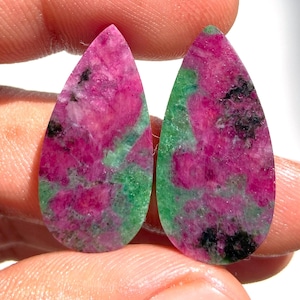 Könnte beinhalten: Zwei tropfenförmige Edelsteine mit einer Mischung aus Rosa, Grün und Schwarz. Die Edelsteine werden zwischen den Fingern gehalten, wodurch ihre polierten Oberflächen zur Geltung kommen. Die rosa Bereiche haben ein marmoriertes Aussehen.