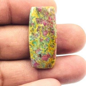 May include: A rectangular gemstone with a mottled pattern of pink, yellow, and green. The stone is held in a hand, showcasing its polished surface and unique colour combination. The gemstone's dimensions are not visible.