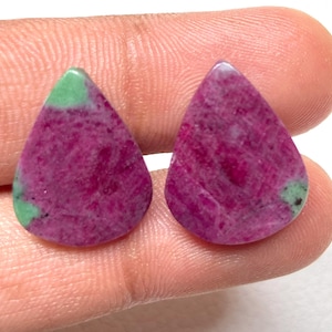 Könnte beinhalten: Zwei tropfenförmige Edelsteine in tiefem Magenta mit grünen Akzenten. Die Steine haben ein strukturiertes Aussehen mit einer Mischung aus Farben und Mustern. Die Edelsteine werden in einer Hand gehalten, um ihre Größe und Form zu zeigen.