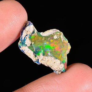 May include: A vibrant, raw opal gemstone with a spectrum of colors including green, red, and yellow. The opal is held between fingers, showcasing its iridescent play-of-color against a dark background. The stone has a rough, natural texture.