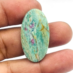 以下が含まれることがあります： さまざまな色合いの楕円形の宝石。石は、黄色の縞模様とピンクと青のパッチがある、明るい緑色のベースが特徴です。宝石は指の間に挟まれています。