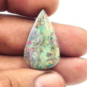 Może przedstawiać: Kształtny kamień szlachetny w kształcie łzy z mieszanką zielonych, różowych i brązowych kolorów. Wypolerowany kamień ma charakterystyczny wzór. Kamień szlachetny jest trzymany w dłoni, co podkreśla jego rozmiar i kształt. Idealny do biżuterii.