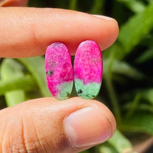 Può includere: Due pietre preziose ovali, ciascuna con una sorprendente sfumatura di colore. La parte superiore è di un rosa brillante, che sfuma in una base verde chiaro. Piccole inclusioni nere sono visibili all'interno dell'area rosa. Le pietre sono tenute tra le dita.