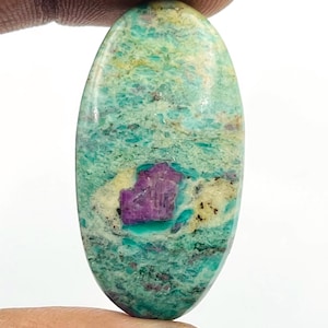 Pode incluir: Uma pedra preciosa oval com uma mistura de cores. A pedra apresenta uma base de verde-azulado e verde com manchas amarelas e uma área central de roxo profundo. A pedra é segurada entre dois dedos.