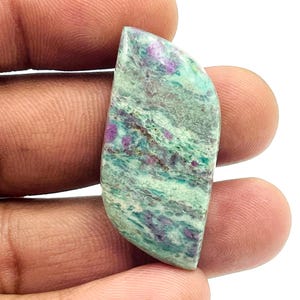 Pode incluir: Uma pedra preciosa polida com um padrão único de tons verdes, azuis e roxos. A pedra tem uma forma lisa e curva e é segurada em uma mão contra um fundo branco. As cores se misturam, criando um efeito marmorizado.