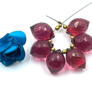 May include: A strand of faceted, deep red gemstone beads with gold-colored spacers. A blue fabric flower is to the left. The beads are strung on a wire, and the background is white. The beads are a deep, transparent red.