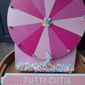 May include: A pink and purple prize wheel with a floral embellishment in the center. The wheel has a white arrow and is mounted on a pink base. The sign below reads "TUTTI CUTIE MYSTERY BUNDLES".
