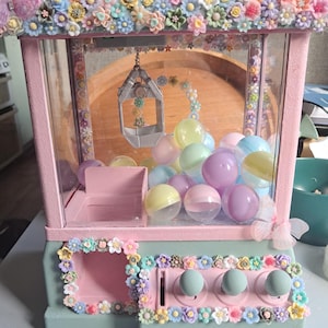 Puede incluir: Una máquina de garras de colores pastel decorada con una abundancia de flores en miniatura. La máquina presenta un marco rosa, una base verde menta y paneles de vidrio transparente. En el interior, hay bolas de plástico de colores. La parte superior está cubierta de pequeñas flores coloridas.