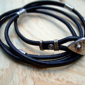 Könnte beinhalten: Ein schwarzes Lederarmband mit silbernen Akzenten. Das Armband hat einen dreieckigen Anhänger mit einer strukturierten Oberfläche.